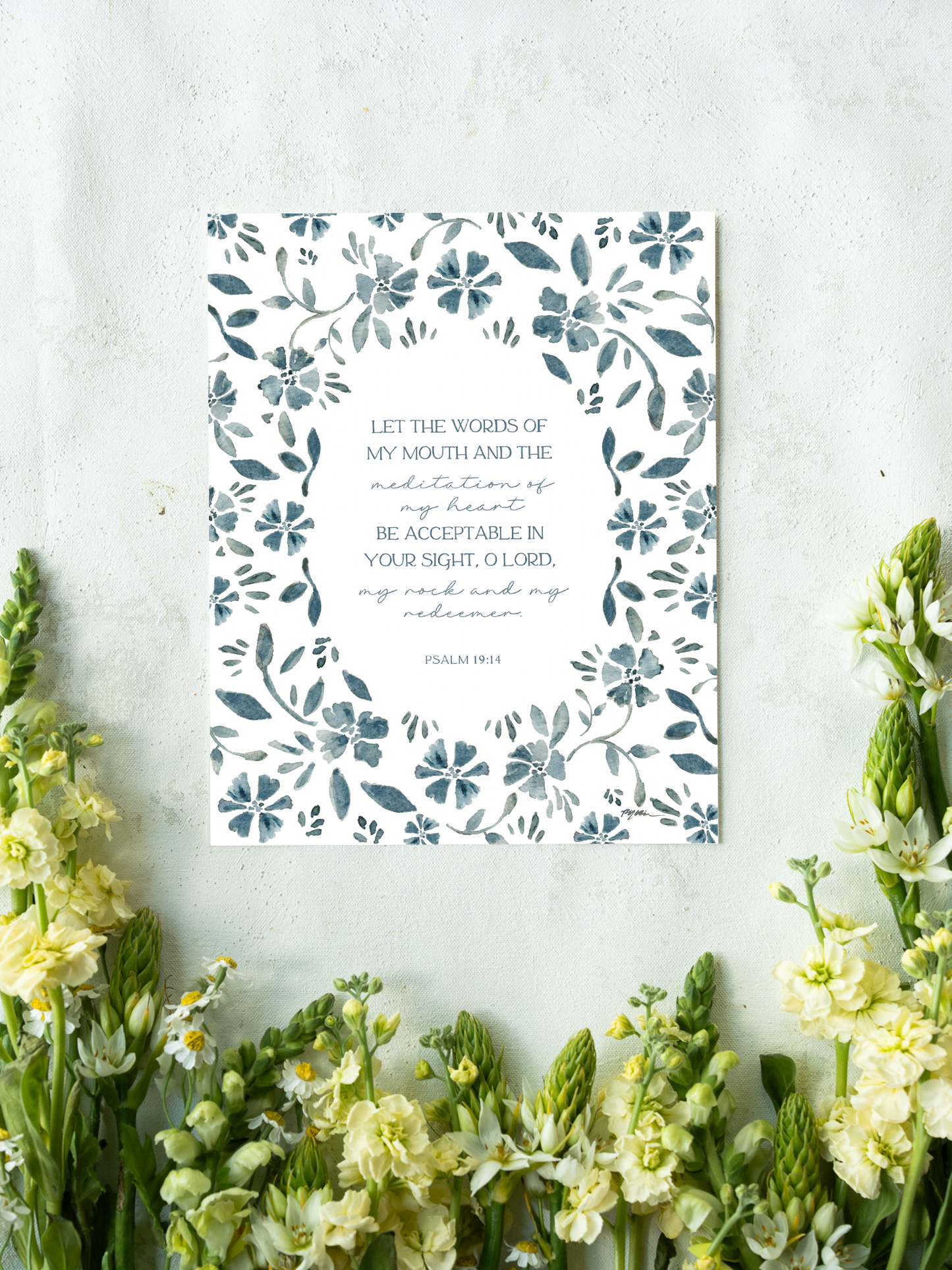 A vertical art print featuring a floral border in slate blue-gray on a white background, with a Bible verse from Psalm 19:14 printed in the center. The print is styled in a flat-lay setting on a textured light gray surface, framed by yellow and white flowers at the bottom.