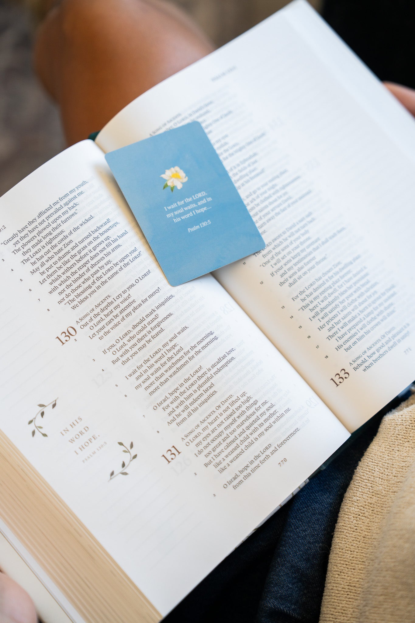 A light blue bookmark card with a small yellow flower illustration and the text 'The Lord's Prayer' lies on an open book held by someone.