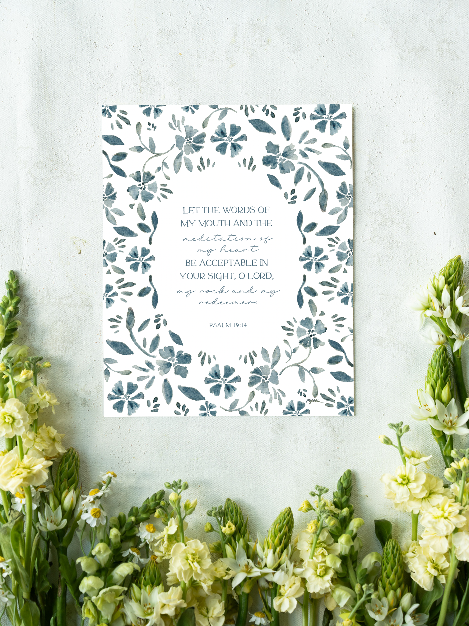 A vertical art print featuring a floral border in slate blue-gray on a white background, with a Bible verse from Psalm 19:14 printed in the center. The print is styled in a flat-lay setting on a textured light gray surface, framed by yellow and white flowers at the bottom.