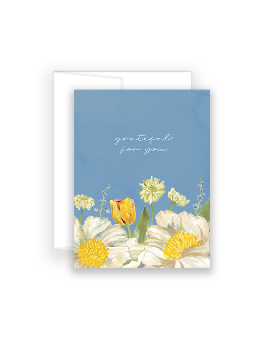 A greeting card with a blue background and watercolor-style white and yellow flowers along the bottom, with the words 'grateful for you' in white script near the top. A white envelope is partially visible behind the card on a light gray background.