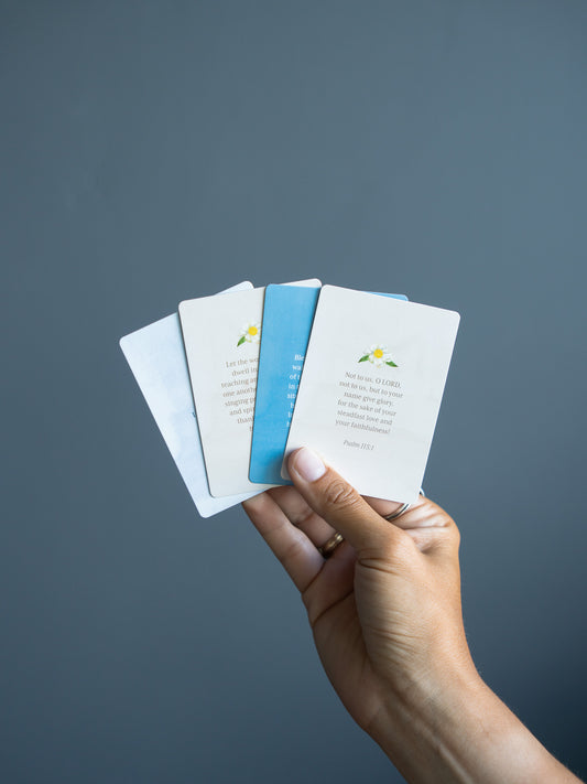 A hand holding a fanned-out set of small printed cards against a dark blue-gray background. The cards are mostly white with light blue and pale green accents, and one card shows a small yellow flower illustration.