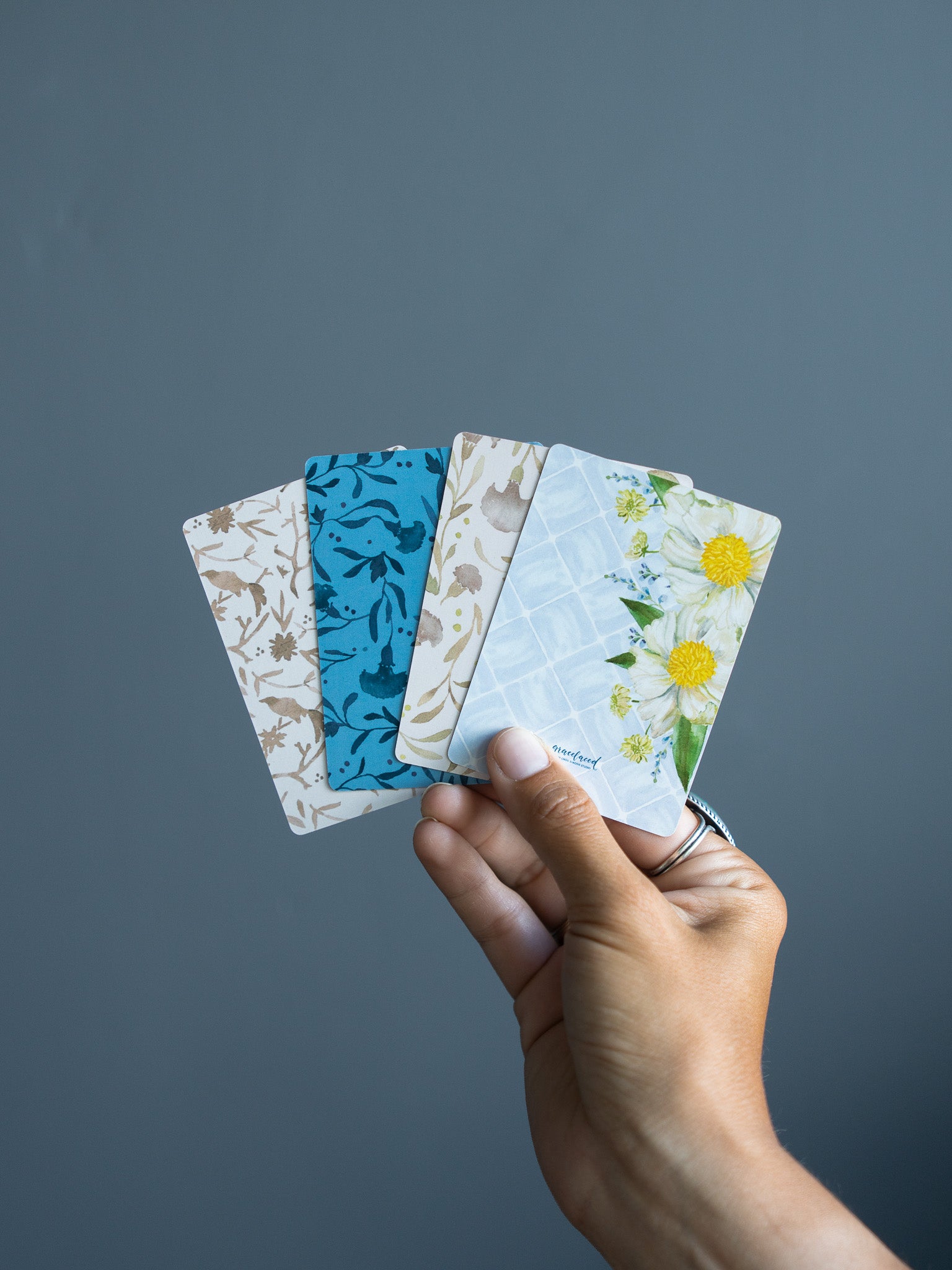 A hand holding four patterned gift cards against a solid dark gray background. The cards feature various designs including blue floral, beige botanical, and a light blue dandelion pattern.