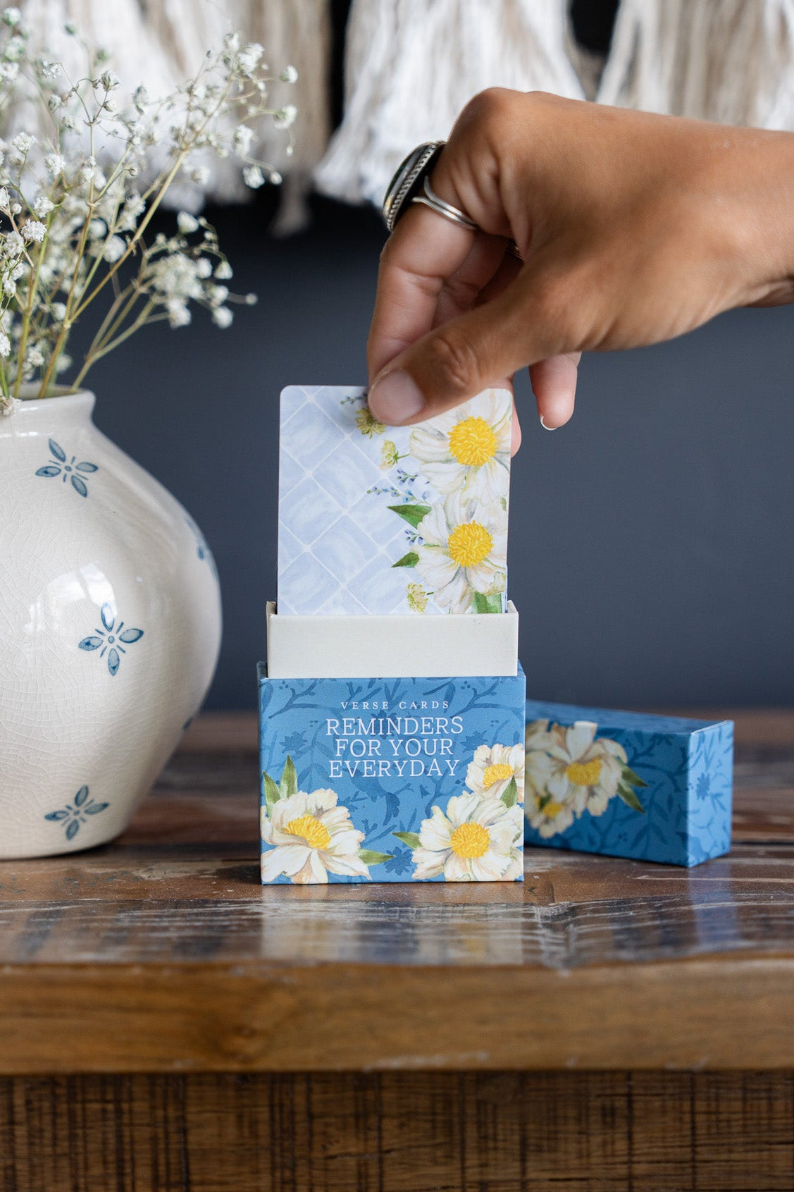 A hand inserts a card into a small blue floral display box on a rustic wooden table, with a white vase of baby's breath in the background.