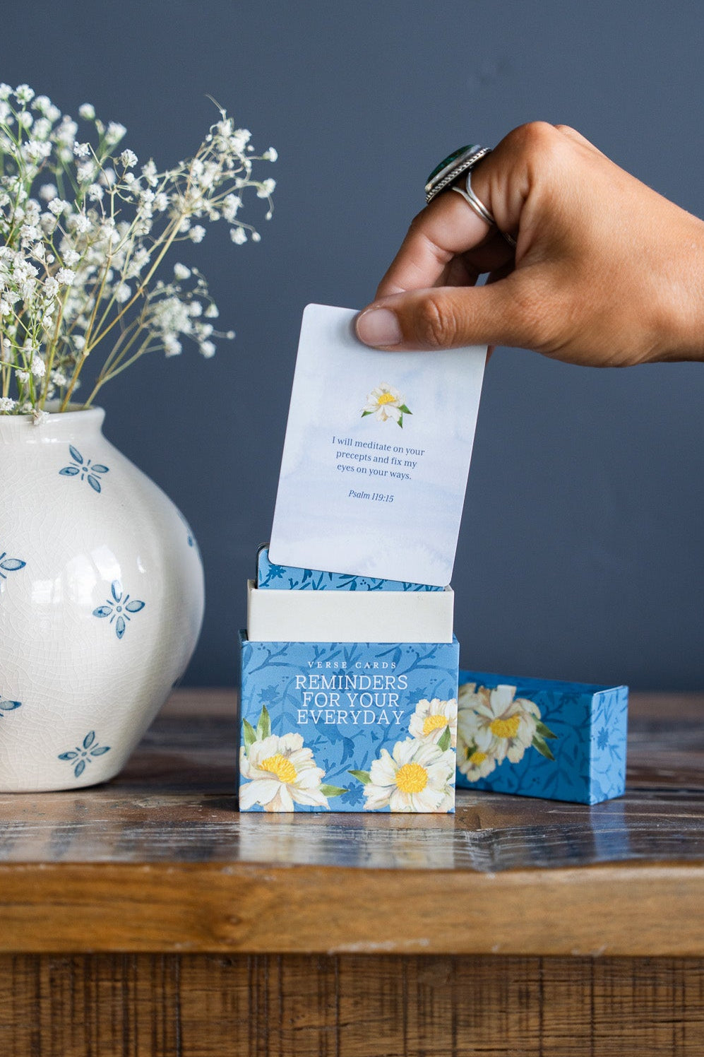 A hand holding a small white card above a blue floral box on a wooden table, with a white ceramic vase of baby's breath to the left against a dark gray wall.