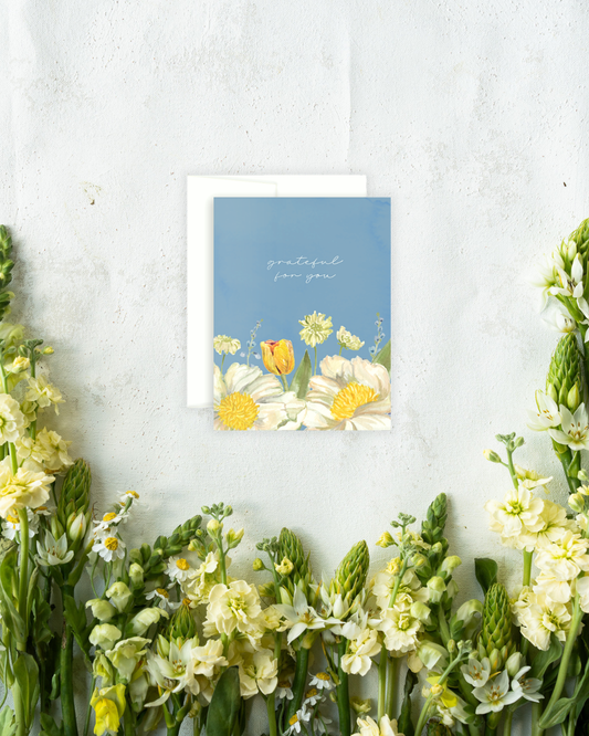 A floral greeting card with a blue background and white script text, placed on a white envelope against a textured white wall, surrounded by yellow and white flowers.