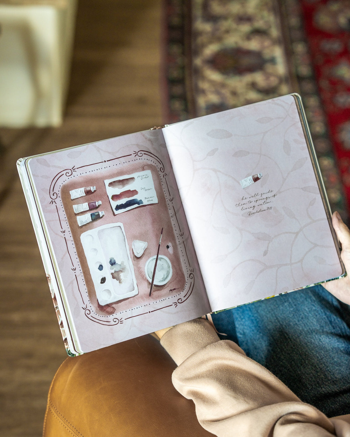An open book held by a person, showing a decorative interior page with a painted palette and small illustrations. The person is wearing a beige top and blue jeans, with a brown leather chair and patterned rugs in the background.