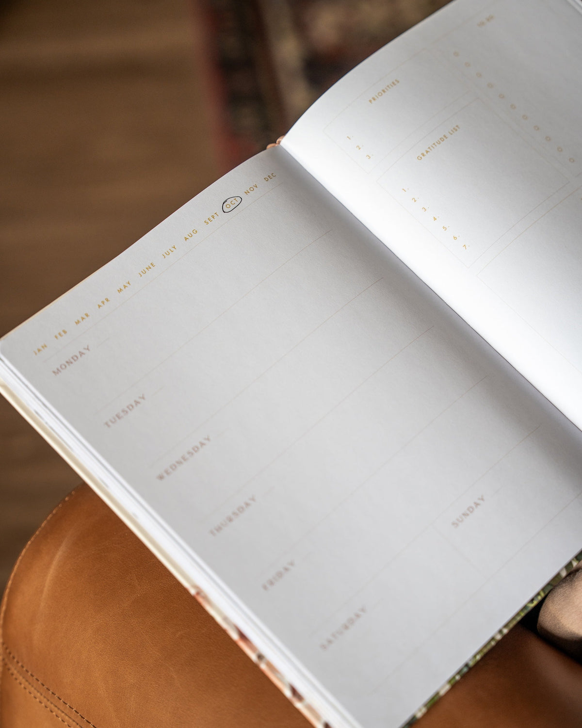 An open weekly planner notebook with blank white pages and gold foil weekday headings, resting on a brown leather surface with a blurred background.