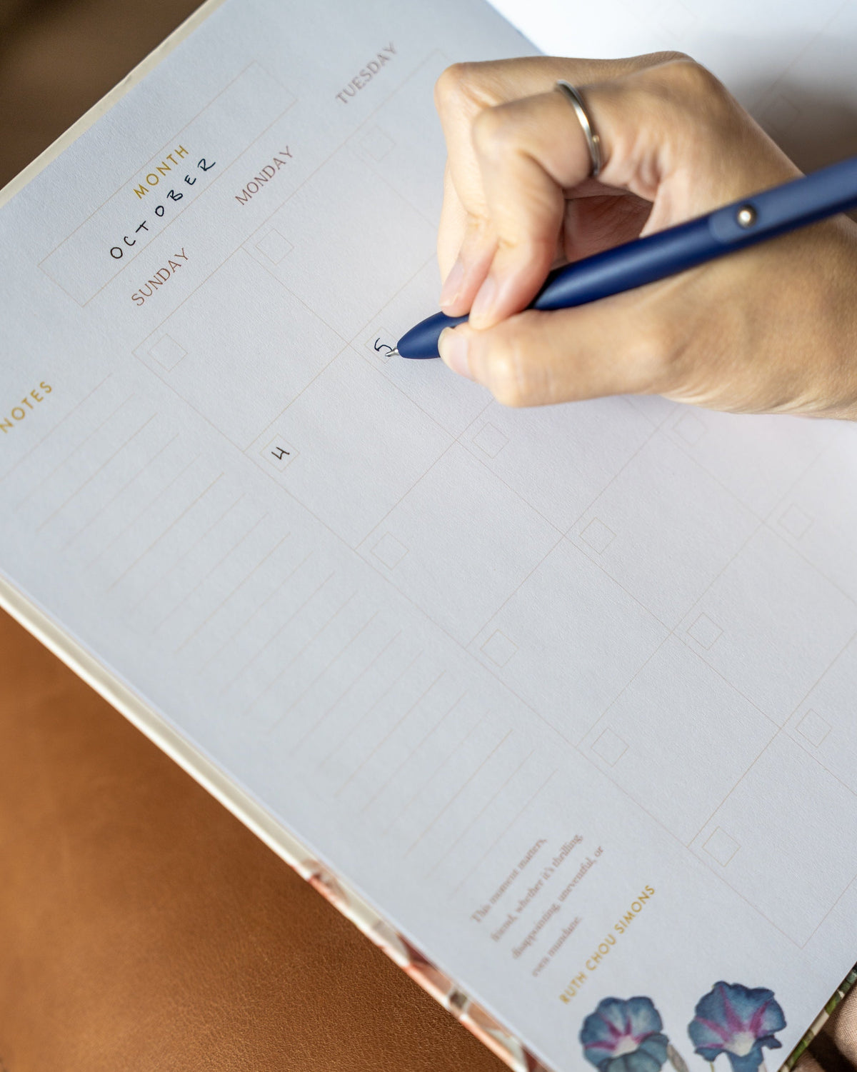 A close-up of a hand writing in a weekly planner pad with a blue pen, resting on a brown leather surface.