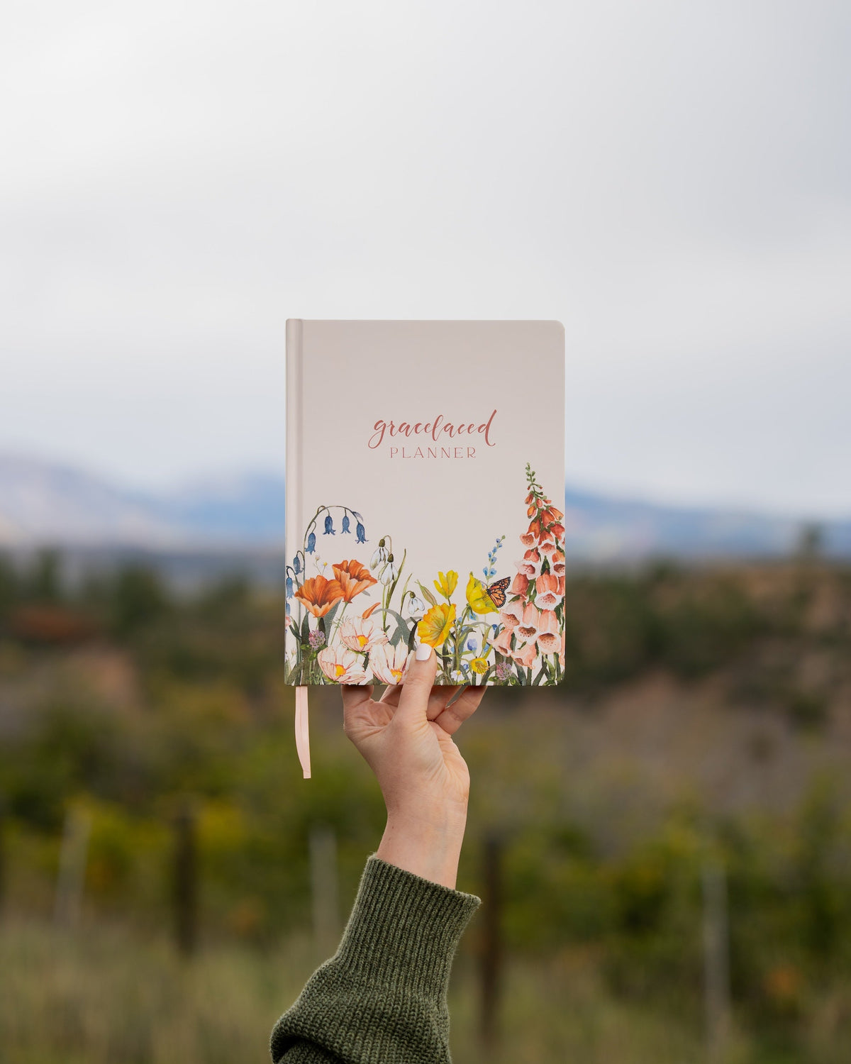 A person holds up a 'graceland planner' with a floral cover design against a blurred outdoor background of trees and mountains.