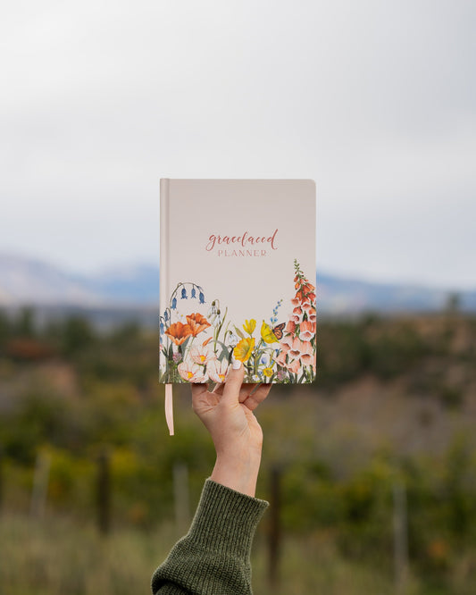 A person holds up a 'graceland planner' with a floral cover design against a blurred outdoor background of trees and mountains.