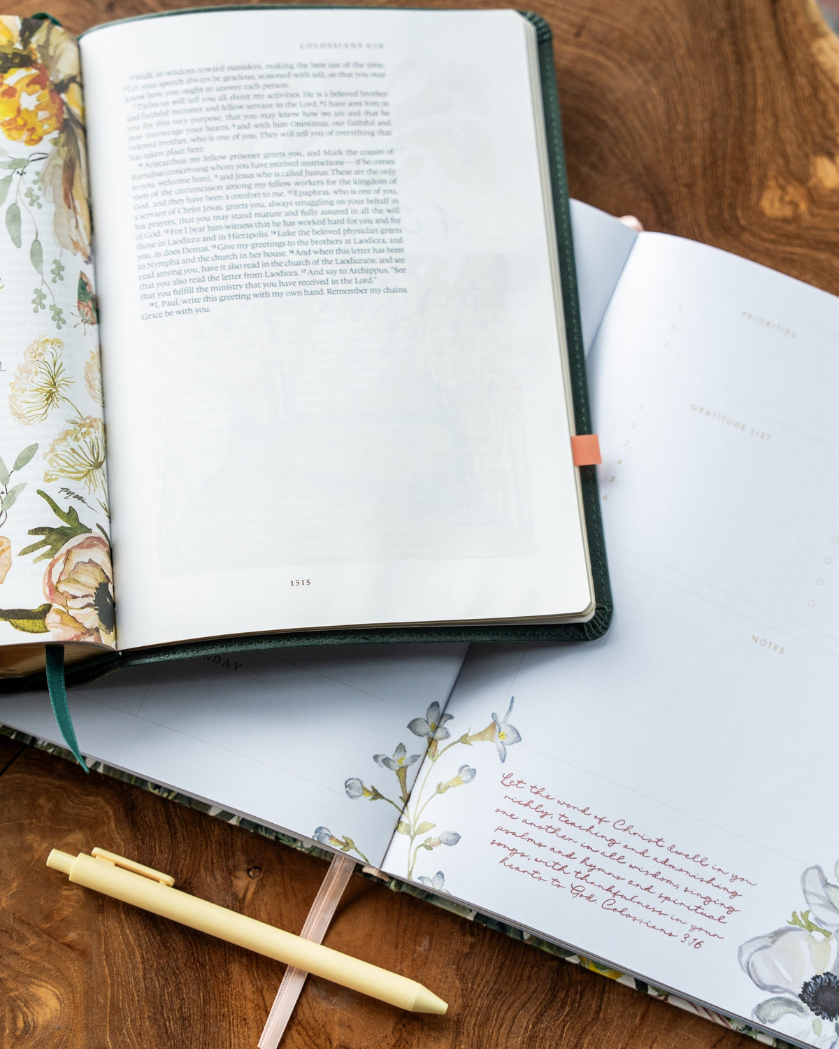 An open floral journal or Bible-style book lies on a wooden surface, with printed text on the left page and decorative floral calligraphy on the right. A pale yellow pen rests on the journal, and another floral-bound book is partially visible underneath.